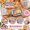 Golden croissant fidget clicker keychain inside a small gray tray-style case with metal keyring